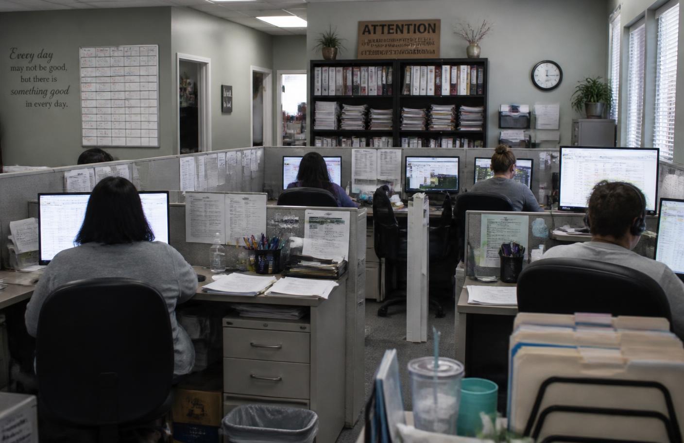 A home-care agency scheduling and billing department with several administrative staff working at desks surrounded by printed schedules and authorization binders.